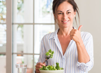 Žena v bílé košili sedí ustolu a jí vidličkou vařenou brokolici. Existují způsoby, jak se poprat se sacharidovou chřipkou.
