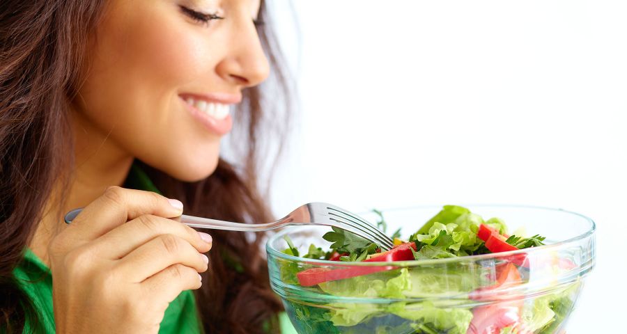 Mladá žena drží skleněnou mísu se zeleninovým salátem, který se chystá ochutnat vidličkou, a usmívá se.